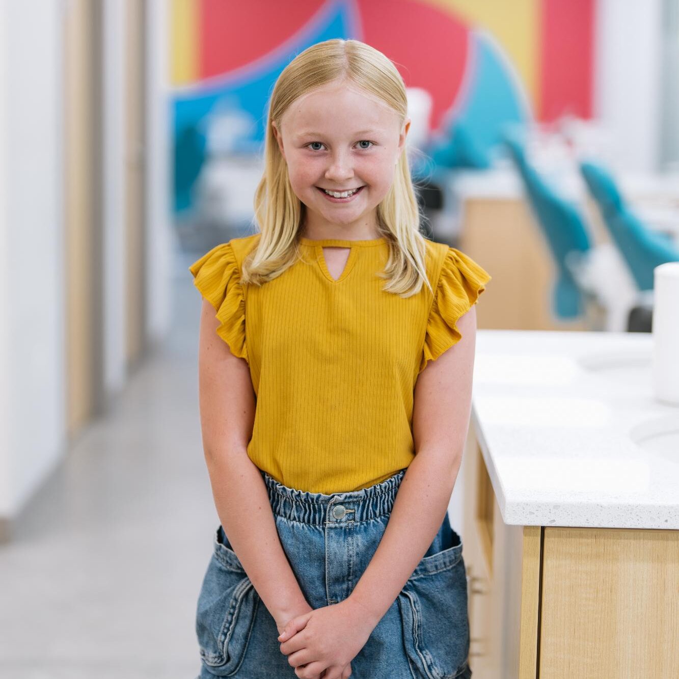 Paradigm-Testimonials-2025-00431 Smiling young girl in a yellow shirt, standing in a dental office, promoting the Puffin Club at Paradigm Orthodontics for children's dental health.